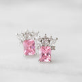 A pair of stud earrings featuring rectangular pink gemstones in silver settings, accented with small clear stones, photographed on a light marble surface.