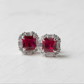 A pair of silver stud earrings featuring square-cut deep pinkish-red center stones surrounded by baguette-cut clear stones, photographed on a light gray surface.