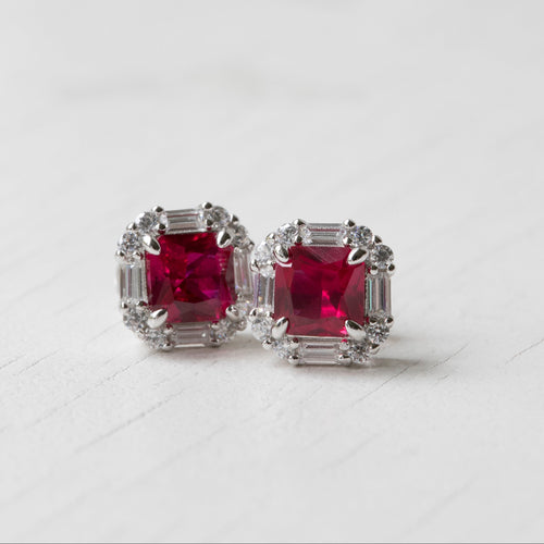 A pair of silver stud earrings featuring square-cut deep pinkish-red center stones surrounded by baguette-cut clear stones, photographed on a light gray surface.