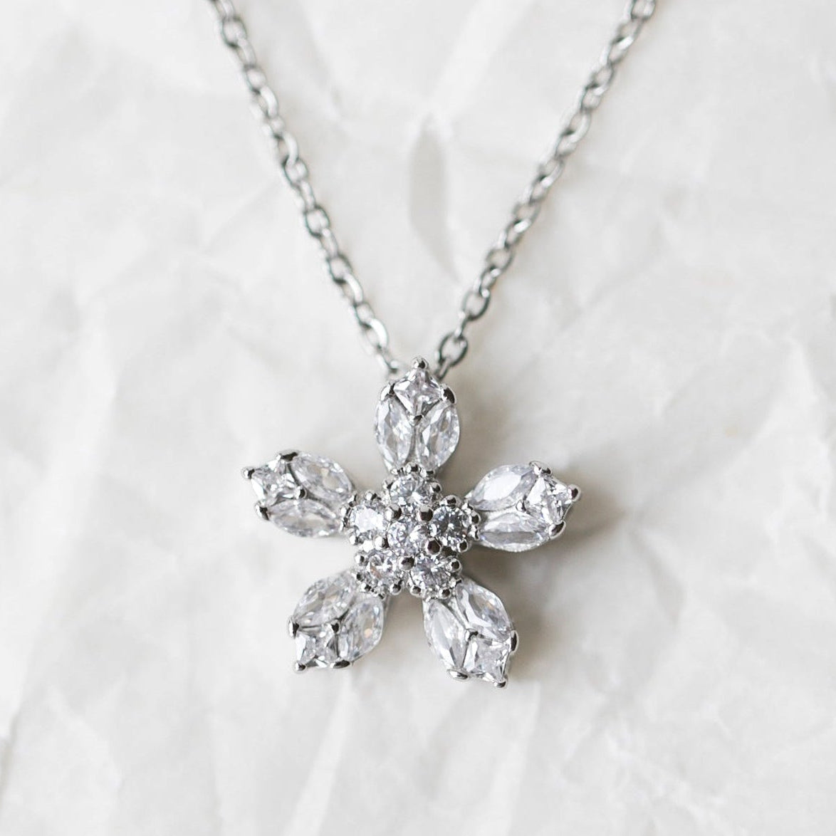 A silver-tone chain necklace with a five-petal flower pendant made of clear marquise-cut, displayed on a textured white background.