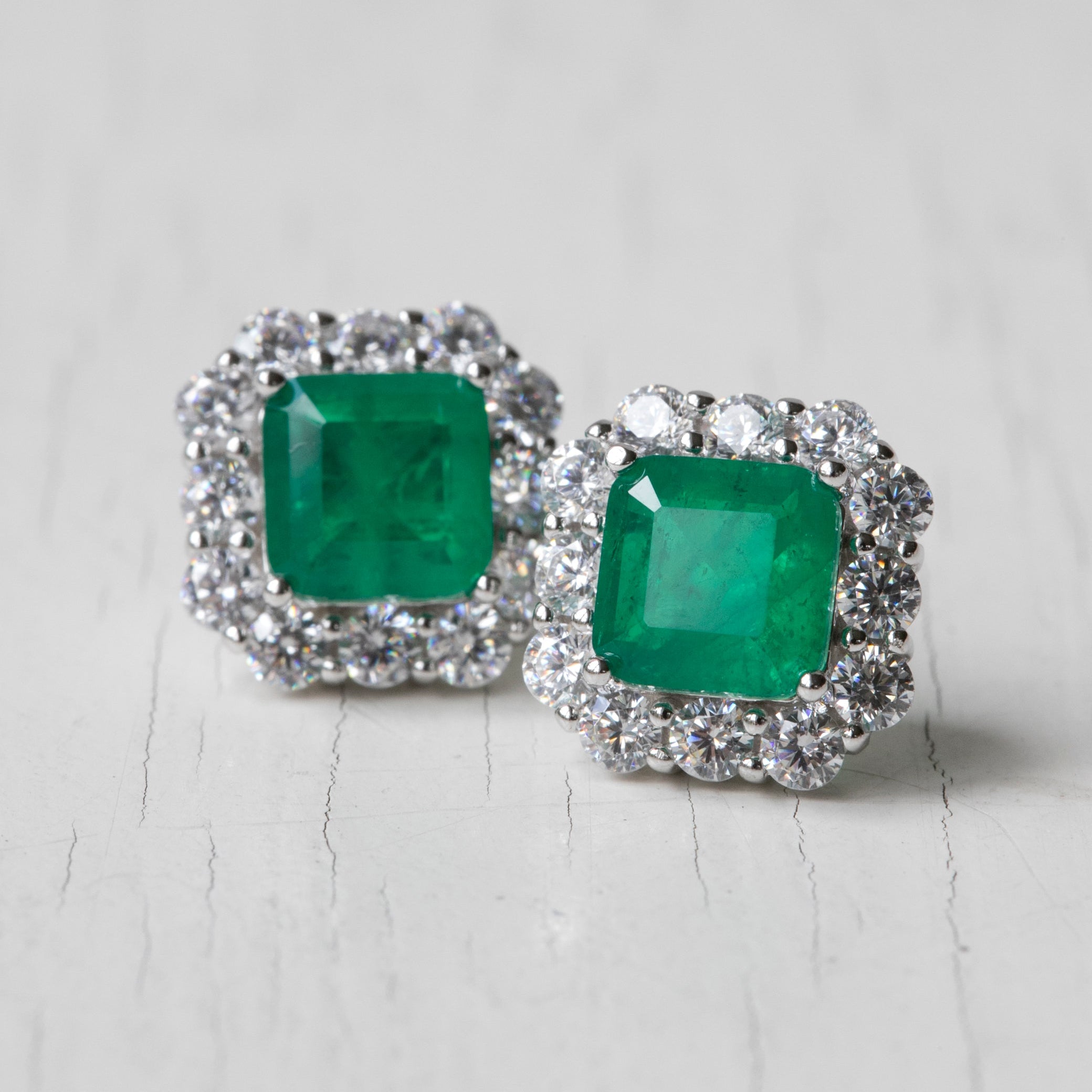 A pair of square stud earrings featuring vibrant green emerald-cut gemstones surrounded by a halo of small clear stones in a silver-toned setting, displayed on a light gray textured surface.