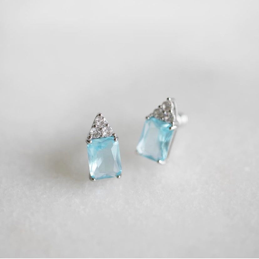A pair of silver stud earrings featuring rectangular light blue gemstones and triangular clear crystal clusters at the top, set against a soft, light gray background.